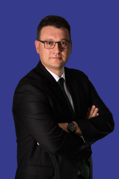 Thomas Christ Ein Mann mit Brille in einem schwarzen Anzug, Arme vor der Brust verschränkt, vor blauem Hintergrund.